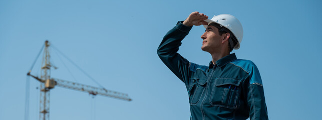 A builder in work clothes and a helmet stands on a construction site against the background of a construction crane. 