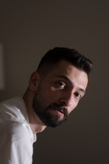Obraz premium Dramatic lighting from an open window illuminates a handsome young man seated in a dark man. He is Syrian, and has serious eyes and a dark beard.