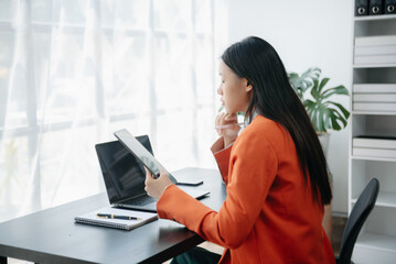  Beautiful Asian woman using laptop and tablet while sitting at her working place. Concentrated at work.