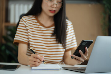 Woman hand working at a smartphone, computer and writing on a notepad with a pen in the home office.