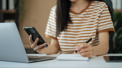 Woman hand working at a smartphone, computer and writing on a notepad with a pen in the home office.