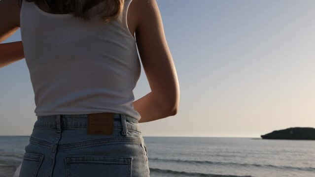 Beautiful young girl taking off her white shirt while standing on the beach looking at the sea. back shot of pretty lady on vacation looking to the horizon on a sunny perfect day and go for a swim.