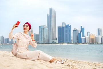girl smiles radiantly at the smartphone camera, wearing red turban, as she poses confidently against the backdrop of the bustling cityscape and tranquil beach.