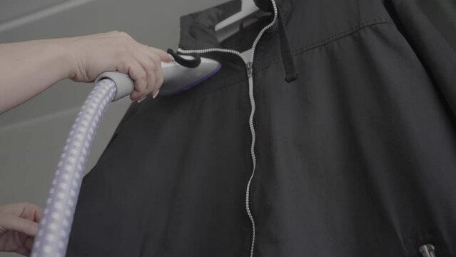 Person uses steam iron to prepare black sports jacket for walking in park. Human irons comfortable walking jacket with steam