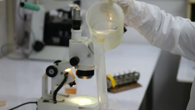 A scientist in a white coat and gloves pours water from a jug into a test tube