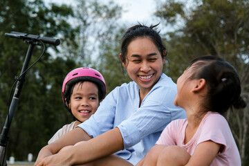 Mother and children sit and rest after riding scooters in the park. Mother and daughters spend their free time riding scooters outdoors together. Happy Loving Family.