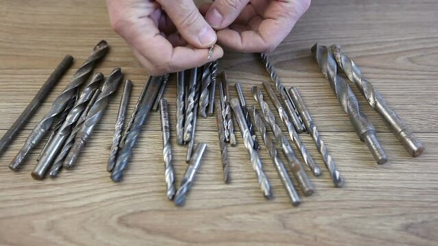 A man chooses a thin drill from the many laid out on the table. Drills for metal and wood are on the table.