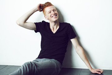 Portrait, ginger and man sitting on a floor with hand on head or relax or smile for fashion and against white wall. Happy, unique style and excited male model with red hair laugh for fun in studio