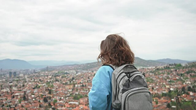A young Caucasian woman with a backpack taking a picture of the cityscape of Sarajevo with her phone.