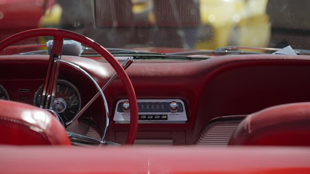Retro dashboard of old vintage convertible with radio and red leather interior and shiny chrome details. Opposite cabriolet is yellow modern muscle car on sunny day. Near cars walk people.