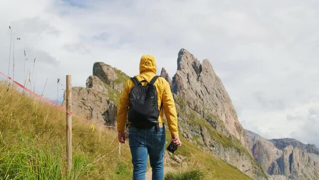 Man holds camera in hand walking toward rocky peak of Seceda mountain. Traveler with backpack enjoys hiking in Italian Alps backside view slow motion