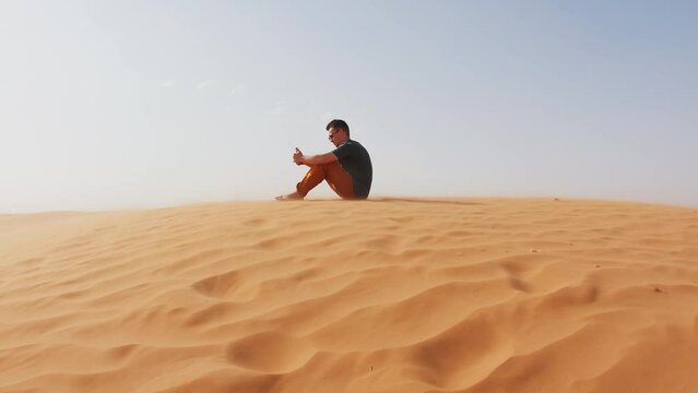 Young man sits in thought on hill in the desert. Concept of loneliness and reflection. Slow motion