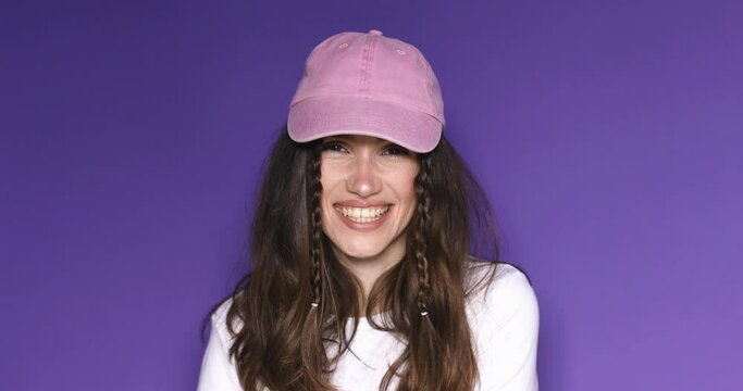 Lovely coquettish brunette woman, winking playfully, flirting, put on pink cap. Woman fix her hair, touch cap isolated on purple background. Curly hair fashion model with two braids face smiling.