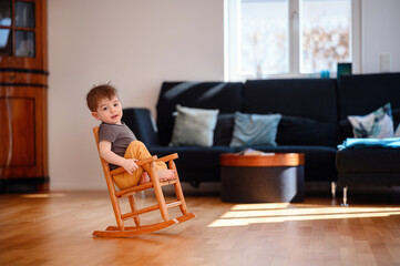 Little toddler boy sitting on wooden rocking chair in living room with dark sofa