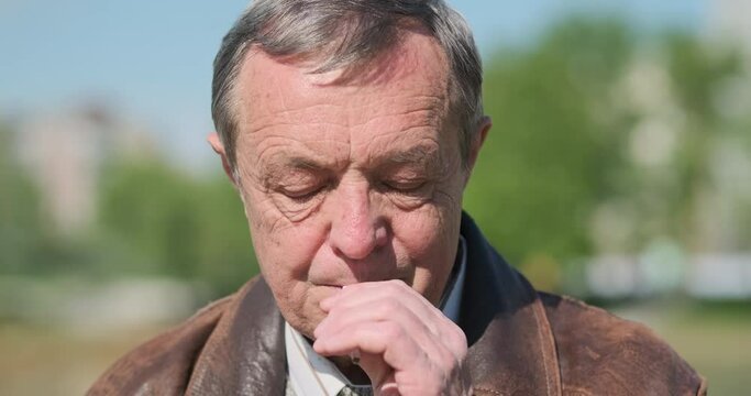 Sad elderly man lights cigarette. With pleasure drags nicotine, releases smoke. City park, portrait, close-up, daytime.