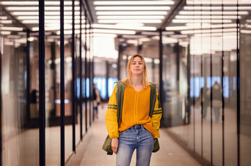 Stylish young blonde girl in yellow green raincoat stands in long corridor between glass showcases