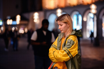 Young blonde girl in yellow-green raincoat standing in night street with lightened house behind © Oleksii Yemelianov