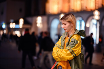 Young blonde girl in yellow-green raincoat standing in night street with lightened house behind