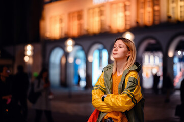 Young blonde girl in yellow-green raincoat standing in night street with lightened house behind
