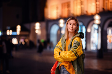 Young blonde girl in yellow-green raincoat standing in night street with lightened house behind