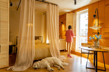 Woman in pink suit opens window shutters, waking up in a sunny and cozy bedroom with a dog sleeping in front