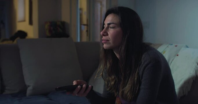 Portrait of a Young Woman Watching TV and Using Remote Control to Choose a Program at Night in her Living Room. Female Enjoying her Alone Time and Relaxing, Channel Surfing, Focusing on the Screen