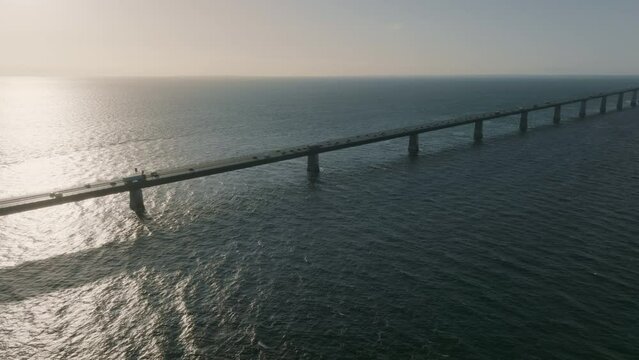 Drone View of Denmark's Great Belt Bridge at sunset, with a roadway stretching majestically over the horizon and connecting two coasts. A flow of traffic moves steadily over the grand structure.