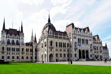 Fototapeta na wymiar The Hungarian Parliament in Budapest. large main dome and smaller towers. arched Gothic windows. fresh bright green lawn. visitors' silhouettes in a distance. famous popular landmark. open square.