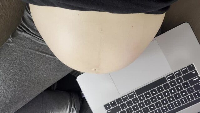 pregnant mom taking a break from working on laptop while breathing comfortably sitting on the couch in lotus position, wearing a black shirt and gray pants with her belly exposed