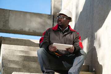 Tired african american worker sits at construction site and holds tablet PC