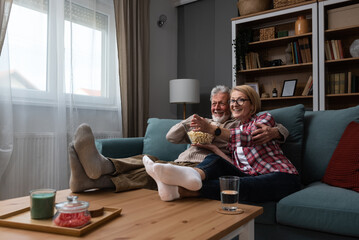 Laughing aged couple, man and woman watching tv, comedy show or movie and eating popcorn snack, sitting on cozy couch at home, mature family, man and woman enjoying free time, weekend together