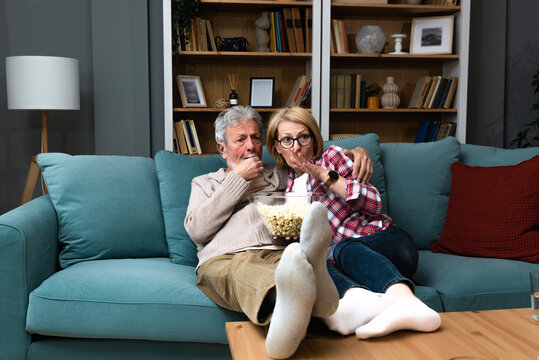 Mature Married Couple Entertaining Observing Terrified TV Program. Senior People Watching Horror Movie At Home With Popcorn Making Scared Faces. Older Husband And Wife Watch Television