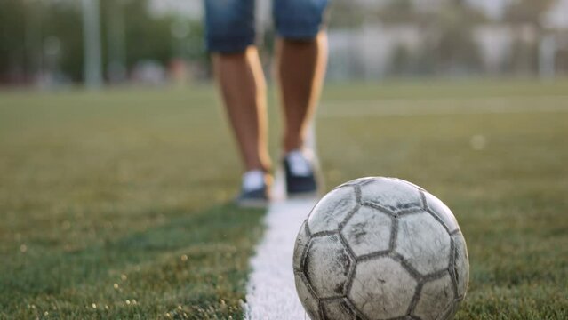 Unrecognizable football player passes old shabby soccer ball, close-up. Ball rolls across football field along white line of penalty area at sunset. Dynamic camera follows rolling sports equipment.