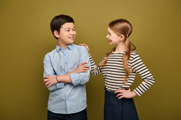 Smiling redhead preteen girl hugging and looking at asian friend folding hands during international child protection day celebration on khaki background