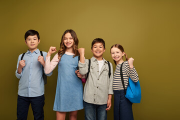 Obraz premium Smiling multiethnic preteen kids with backpacks showing fists as power gesture and looking at camera during world child protection day celebration while standing together on khaki background