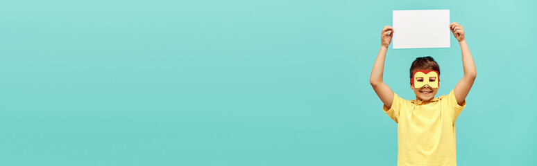 carefree multiracial boy in yellow superhero costume with mask holding blank paper above head on...