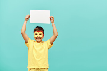 smiling multiracial boy in yellow superhero costume with mask holding blank paper above head on...