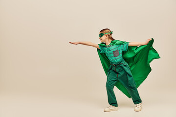 side view of happy kid in superhero costume and mask holding green cloak and standing with...
