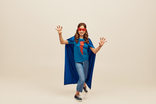 joyful girl in superhero costume with blue cloak and red mask on face, showing hand gesture, standing in denim jeans and t-shirt while celebrating Child protection day holiday on grey background