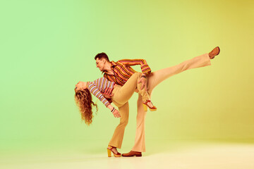 Romantic look. Beautiful young couple, man and woman in vintage stylish clothes dancing disco dance against gradient green yellow background. Concept of retro style, fashion, art, hobby, music, 70s