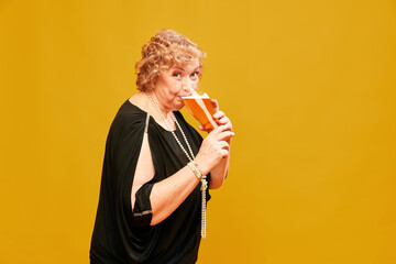 Portrait with senior lady, woman wearing black vintage dress holding big beer mug and drinking with pleasure over yellow background