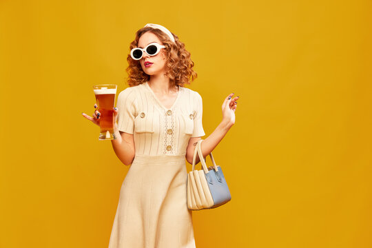 Young beautiful girl wearing stylish retro clothes with sunglasses holding glass of beer over yellow background. Vintage fashionista