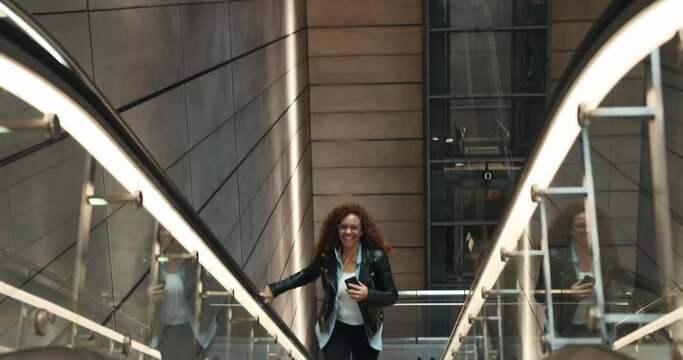 Stylish young woman smiling and listening to music on her cellphone while riding up a metro station escalator 