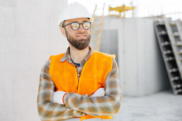 Builder repairman, foreman in safety helmet and vest stands at his workplace in a building under construction with his arms crossed in front of him