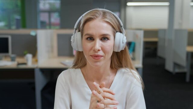Portrait of a cheerful woman designer against the background of international office space, young woman in headphones communicates by video call, talks looking at the camera, template.