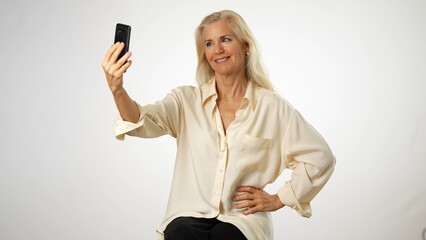 Beautiful mature woman taking selfie with smartphone. Attractive middle aged blond gesturing while looking at phone. Concept of self portrait on white background