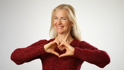 Cheerful happy senior mature woman shows heart gesture with hands showing love, smiling isolated on solid white background