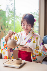 Young woman wearing traditional kimono sitting in a Japanese style house pouring tea to drink, garden background