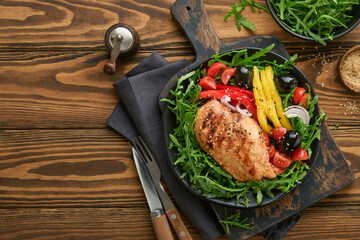 Salad with grilled chicken breas fillet with fresh arugula, tomatoes and olives on black plate on old wooden dark background. Chicken meat with salad. Ketogenic lunch. Healthy food. Top view.