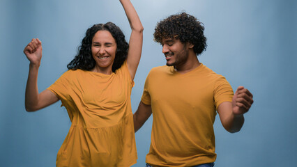 Funny excited happy couple Hispanic woman Latino wife girlfriend dance with carefree Indian man active cheerful husband boyfriend dancing having fun celebrate fooling around at blue studio background
